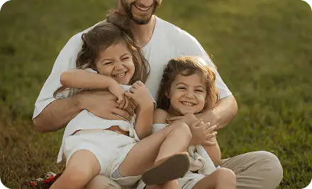 A father sitting on grass hugging his two laughing daughters