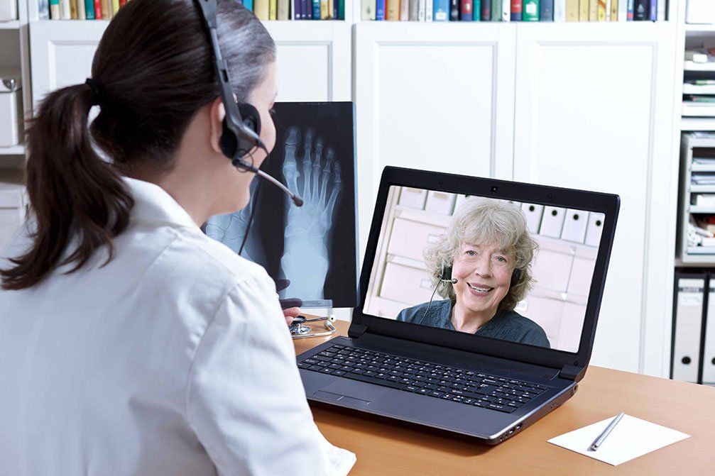 A clinician wearing a headset holds an X-ray while video-calling an older patient on a laptop